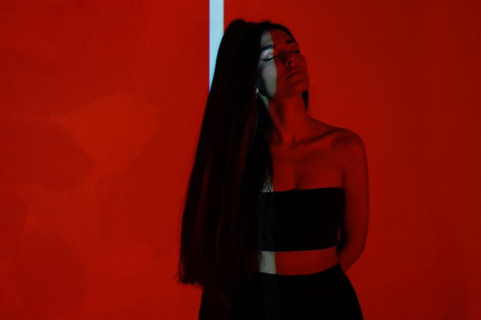 A woman with long dark hair stands against a vivid red background wearing a black strapless top, bathed in moody lighting.
