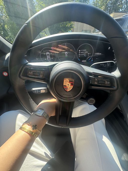 A first-person view from the driver's seat of a Porsche, showing hands wearing a watch and bracelets gripping the steering wheel, with the digital dashboard displaying a speed of 0 mph.