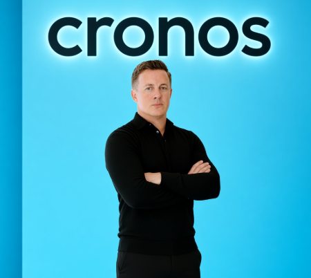 A confident man in a black polo shirt stands with his arms crossed against a bright blue backdrop featuring the bold "cronos" logo. The image appears to be a promotional or brand-style shot for Cronos (CRO), the cryptocurrency referenced in the post speculating about its price returning to $0.05 by year's end.