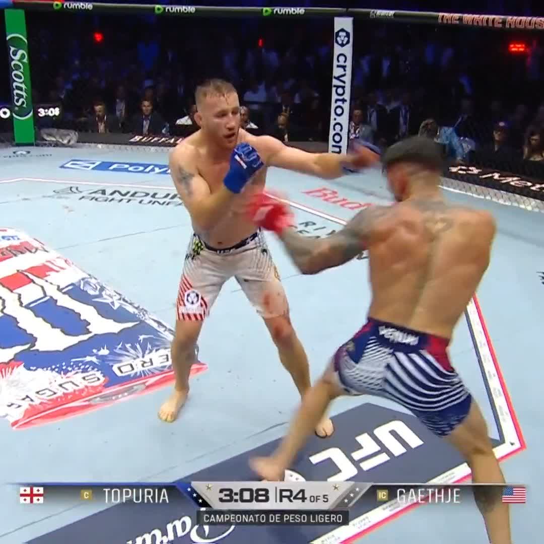 The image shows two boxers in a ring, one wearing white shorts and the other wearing blue shorts, both throwing punches at each other during a fight. The fighter with the white shorts has his fist raised high as he prepares for impact. The fighters are surrounded by an audience of people watching the match intently from various positions around the ring.