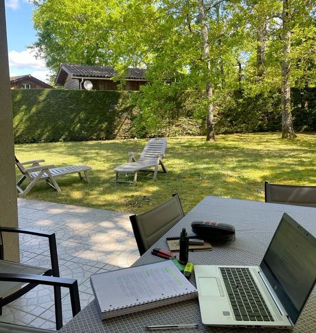 The image shows a laptop computer sitting on top of a table, with a notebook or planner next to it. The scene takes place outdoors, possibly at an outdoor cafe or patio area. There are two chairs nearby, one closer to the camera and another further away. A person can be seen in the background, but their face is not visible. The image also contains text that reads "Focus on what is in the image and how it relates to the post."