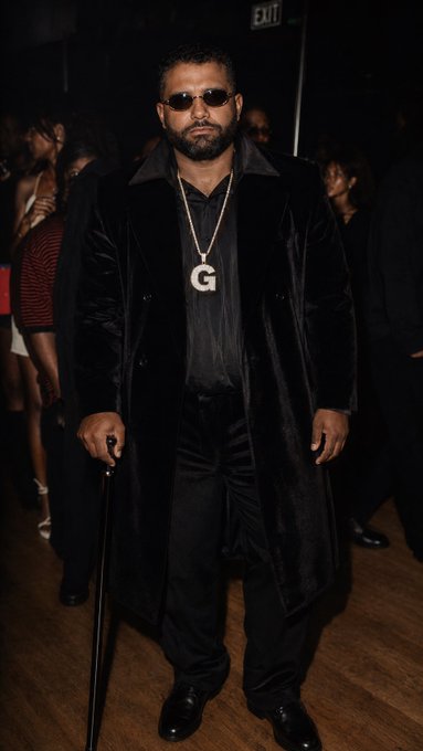 A man wearing dark sunglasses and a long black coat stands holding a cane, accessorized with a prominent chain featuring a 'G' pendant.