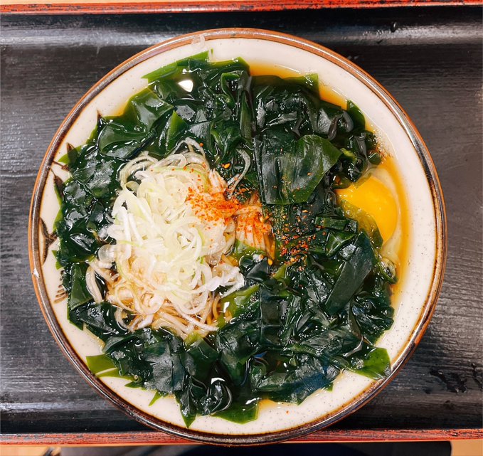 A bowl of ramen or noodle soup topped with wakame seaweed, shredded white onions, a raw egg yolk, and chili powder, served in a traditional ceramic bowl.