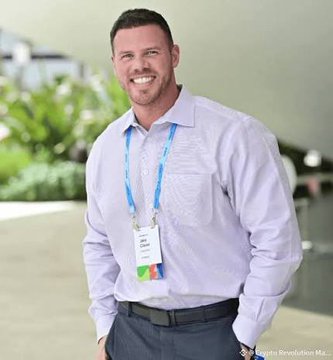 A smiling man in a light blue button-down shirt wears a lanyard with a credential badge in what appears to be a conference or event setting. The image serves as a professional portrait to accompany the post's quote from Jake Claver about patience in holding XRP while others chase short-term gains.