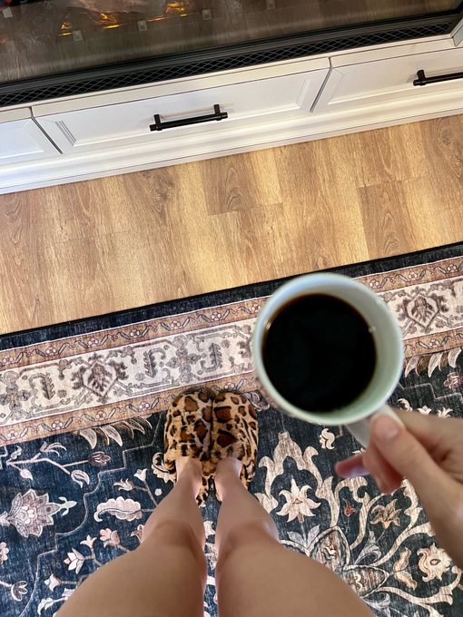 A woman in leopard-print slippers stands on a floral patterned rug while holding a mug of black coffee, embracing a peaceful morning routine that celebrates the simple joy of being alive.