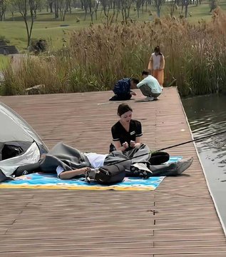 Two people are relaxing on a colorful blanket on a wooden deck by a lake, with one person lying down under a hat while the other sits beside them.