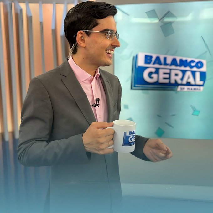 A man in a pink shirt and brown blazer smiles while holding a "BALANCO GERAL" branded mug in a TV studio setting with the show's logo visible in the background.