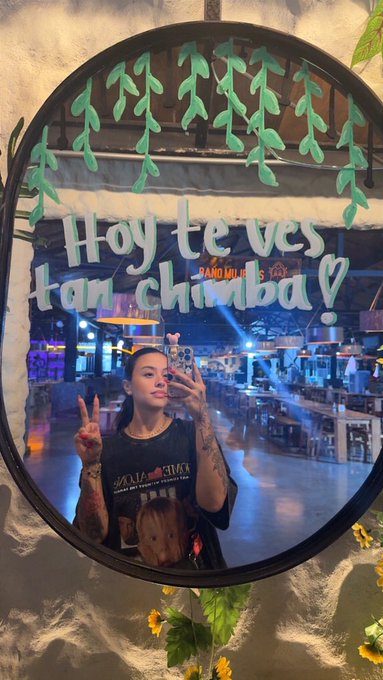 A mirror selfie of a woman with tattoos holding a phone, with decorative text 'Hoy te ves tan chimba' and green leaf decals around the mirror frame.