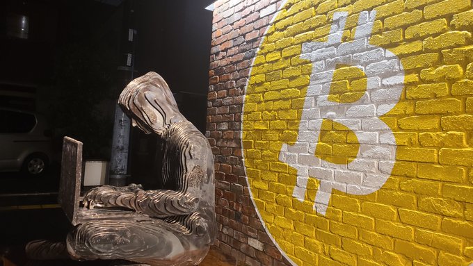 A hooded figure painted to resemble Bitcoin sat at a laptop next to a yellow Bitcoin logo on a brick wall at the Tokyo Bitcoin Base, fitting the Bitcoin-themed study session about the upcoming shift to Japan's Financial Instruments and Exchange Act.