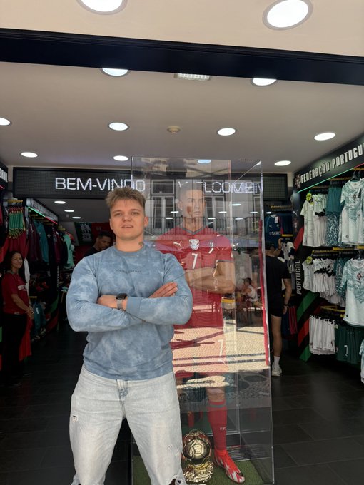 A young man with crossed arms poses in front of a glass display case containing a mannequin wearing a red Portugal #7 Cristiano Ronaldo jersey and a golden soccer ball.