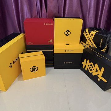 A collection of branded merchandise boxes from Binance and HeyBinance are displayed together, showcasing promotional packaging with the recognizable Binance and HeyBinance logos. The image celebrates Binance's upcoming 9th anniversary, with the caption humorously reflecting on how the user has aged from a wide-eyed university student to a 2002-born "middle-aged" crypto enthusiast since joining the platform.