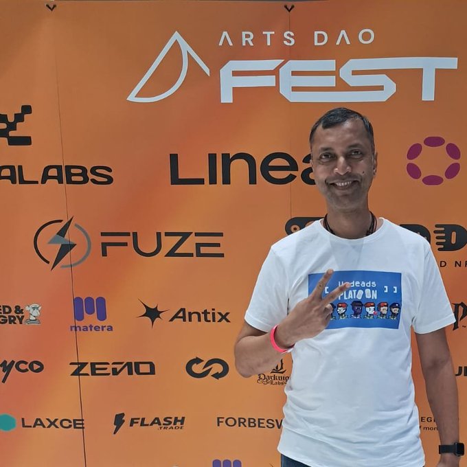 A smiling man stands in front of an orange 'ARTS DAO FEST' sponsor backdrop featuring logos like Linea, Fuze, Antix, and Forbes, making a peace sign while wearing a 'THREADS LATO ON' t-shirt.