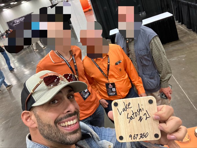 A selfie of a smiling man wearing a cap and sunglasses, holding a wooden paddle that reads "Lake Satosh #1 967360," with three other people in orange shirts blurred in the background.