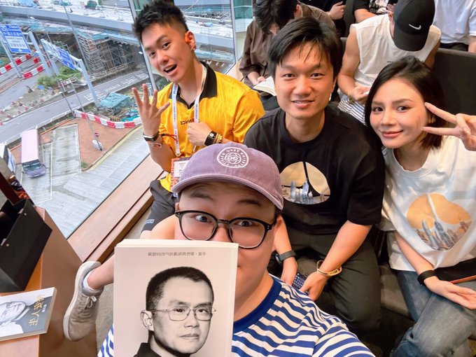 A group selfie of five people at what appears to be a stadium event, with one man in the front holding a book featuring a portrait on the cover.