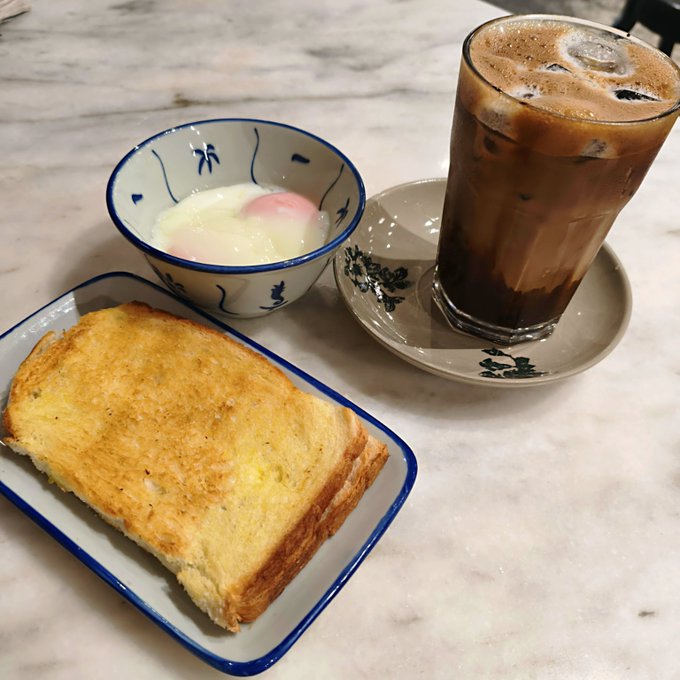 A traditional kaya toast set featuring two slices of golden toasted bread, a bowl of soft-boiled eggs with a pink-and-white yolk, and an iced coffee served in a tall glass on a marble table.

This cozy breakfast spread fits perfectly with the "GM" weekend post, suggesting the 0.0012 ETH treat-yourself moment went to a satisfying, nostalgic Singaporean-style breakfast.