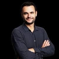 A professional headshot of a smiling man with a beard wearing a dark button-down shirt against a black background.