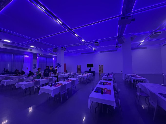 A large room bathed in blue lighting is set up with rows of tables covered in white cloths and chairs, with a few people standing near the back.