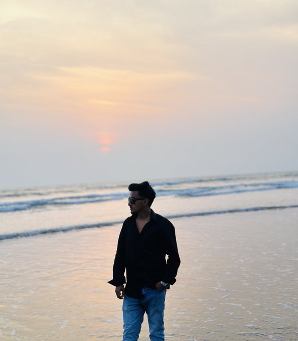 A man in a black shirt and jeans stands on a sandy beach at sunset, looking out toward the ocean.