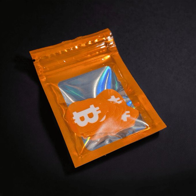 An orange resealable plastic bag lies on a dark surface, containing what appears to be orange Bitcoin-themed tokens or coins with the recognizable 'B' logo.