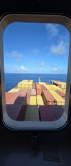 A view through a ship's porthole shows a vast deck stacked with colorful shipping containers stretching toward the horizon under a blue sky, illustrating the massive scale of cargo shipping referenced in the post about delivering packages despite risks of "rugs and drainers."