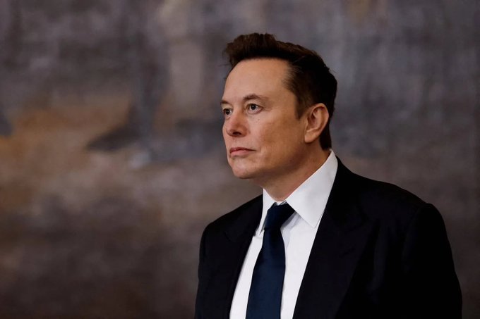 A man resembling Elon Musk, with short brown hair, wears a dark suit, white shirt, and navy tie while looking off to the side against a blurred neutral background.