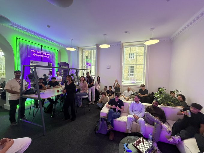 A diverse group of crypto developers gathers in a modern lounge space with purple and green neon lighting, with a screen displaying "Win and flex with @superteamuk." Attendees sit on white couches and stand around a table with drinks, reflecting the post's enthusiasm about connecting with SuperteamUK teams including HawkFi, Otus_Finance, and others during a London meetup.