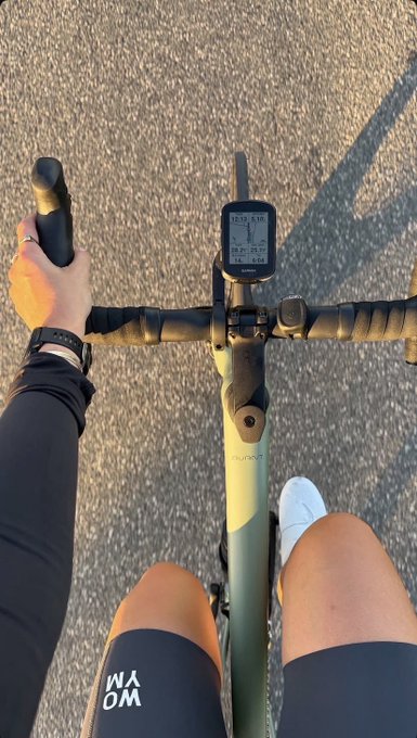 A first-person POV photo of someone cycling, showing their hands on the handlebars of a green bike, a Garmin bike computer, and their legs wearing black compression sleeves with 'WA OM' text and white shoes.
