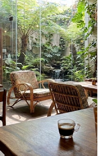 A small glass cup of espresso sits on a wooden table next to wicker chairs, with a lush indoor garden and small waterfall visible through glass in the background.