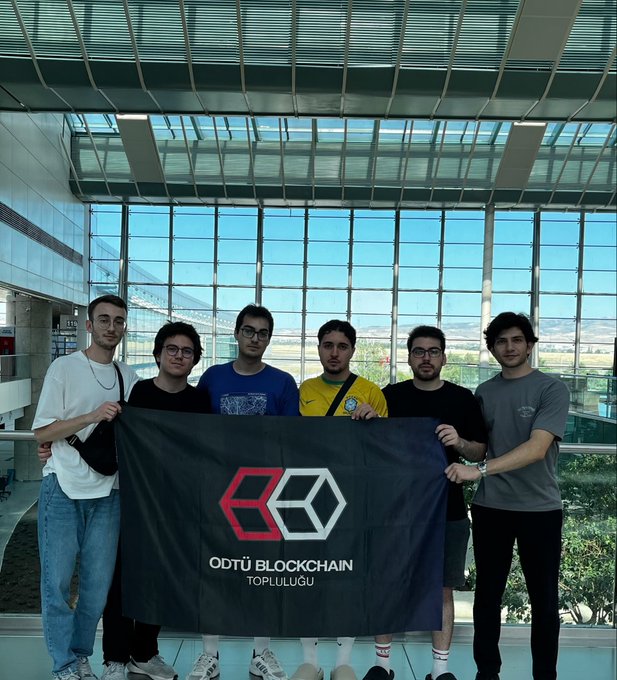 A group of six young men holding a black banner reading 'ODTÜ BLOCKCHAIN TOPLULUĞU' in a modern glass-walled building.