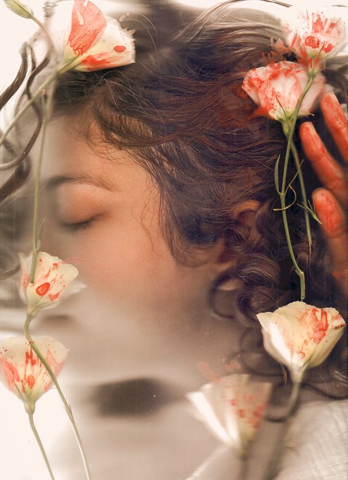 A soft-focus, dreamlike portrait of a young woman with closed eyes, framed by delicate white blossoms stained with red.