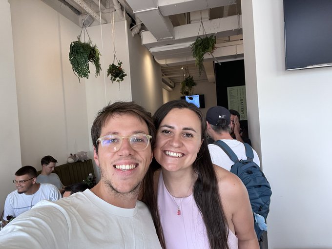 A smiling couple takes a selfie in a bright room with hanging plants, with other patrons visible in the background.