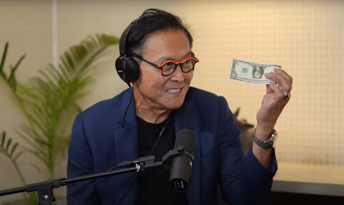 Robert Kiyosaki, author of 'Rich Dad Poor Dad,' wearing headphones and glasses while holding up a US dollar bill during what appears to be a podcast recording.