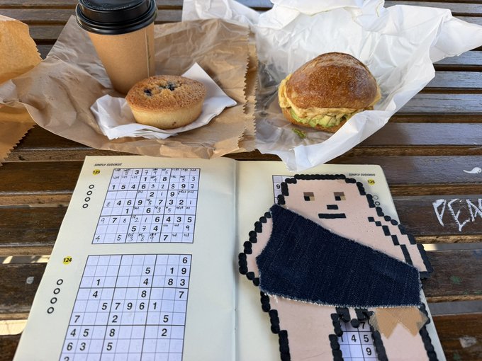 An open puzzle book on a wooden table showing a completed pixel-art puzzle of a character holding a blue object, accompanied by a muffin, sandwich, and coffee cup in the background.