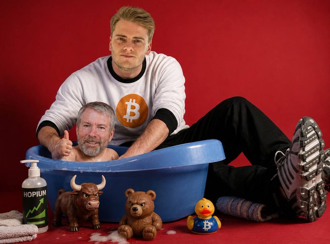 A young man wearing a white Bitcoin logo sweatshirt gives a thumbs-up while playfully bathing a bearded man in a blue plastic tub, surrounded by Bitcoin-themed props including a bull figurine, a teddy bear, a rubber duck with the "B" symbol, and a soap dispenser labeled "HOPIUM." The scene humorously illustrates the post's caption of "giving Saylor his daily hopium bath," referencing Michael Saylor and the optimistic enthusiasm among Bitcoin supporters.