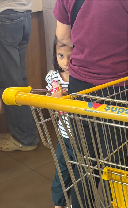 A young child with a serious expression stands behind a yellow shopping cart in what appears to be a supermarket, partially obscured by an adult wearing a maroon shirt.