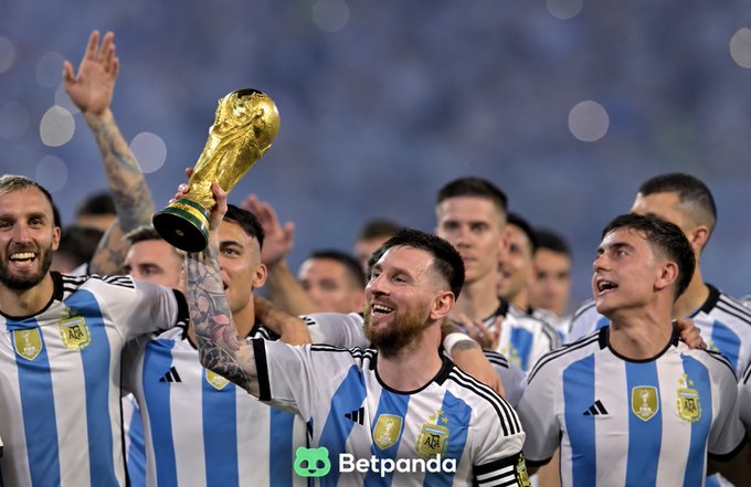 Lionel Messi holding up the FIFA World Cup trophy while celebrating with Argentina teammates in their blue and white striped jerseys, with a Betpanda logo watermark visible.