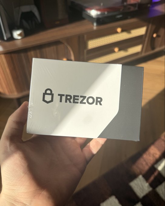 A hand holding a sealed white and gray Trezor hardware wallet box, with a wooden mid-century modern cabinet visible in the background.