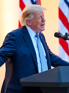 A man with blonde hair wearing a dark blue suit and light blue tie speaks at a podium with American flags behind him, delivering a stern statement regarding potential military action against Iran, with the accompanying news caption highlighting his remarks about choosing between economic devastation or military confrontation.