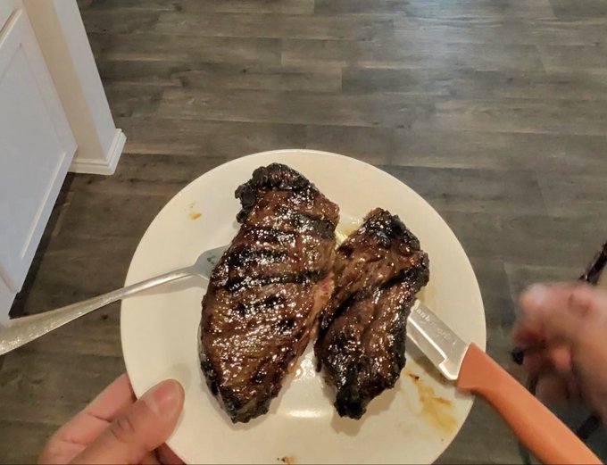 A first-person view of a hand holding a white plate with two pieces of grilled steak, a fork, and a knife with an orange handle, taken over a wooden floor.