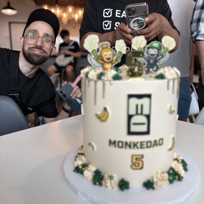 A man wearing a black cap smiles beside a MonkeDAO 5th anniversary cake decorated with banana accents and cartoon monkey toppers, celebrating his birthday alongside the NFT community's milestone.
