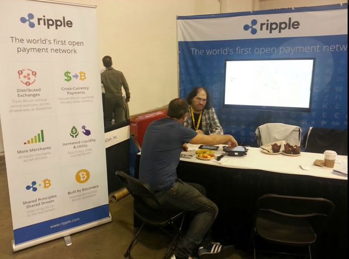 Two men sit at a Ripple booth promoting "the world's first open payment network," featuring banners highlighting distributed exchanges, cross-currency payments, and XRP/Bitcoin branding, as they discuss how a higher XRP price strengthens the XRP Ledger's effectiveness for payments.