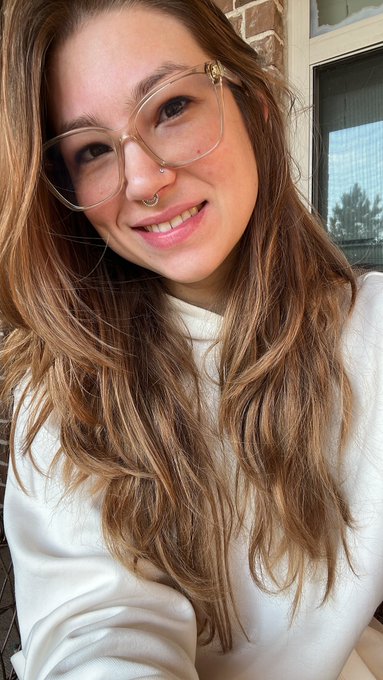 A smiling young woman with long brown hair, clear-framed glasses, and a nose ring takes a selfie wearing a white sweatshirt.
