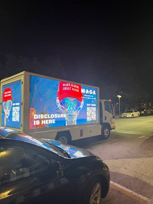 A truck with a digital billboard parked on a street at night. The display features a stylized face with an American flag bandana and text reading "MAKE ALIENS GREAT AGAIN", "MAGA", "DISCLOSURE IS HERE", and "BUY NOW" alongside a QR code.
