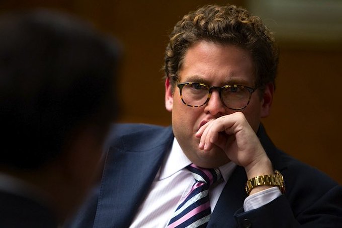 A man with curly hair and tortoiseshell glasses, wearing a navy suit and striped tie, sits thoughtfully with his hand over his mouth in what appears to be a hearing or formal meeting.