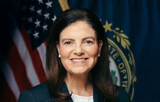 An official portrait of New Hampshire Governor Kelly Ayotte smiling in front of an American flag and the state seal. The image accompanies news about her expressing support for the state to seriously consider Bitcoin adoption.