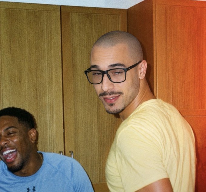 Two men posing for a photo in front of wooden cabinets, with one man wearing glasses and a yellow shirt looking at the camera while another man in a blue Under Armour shirt laughs.
