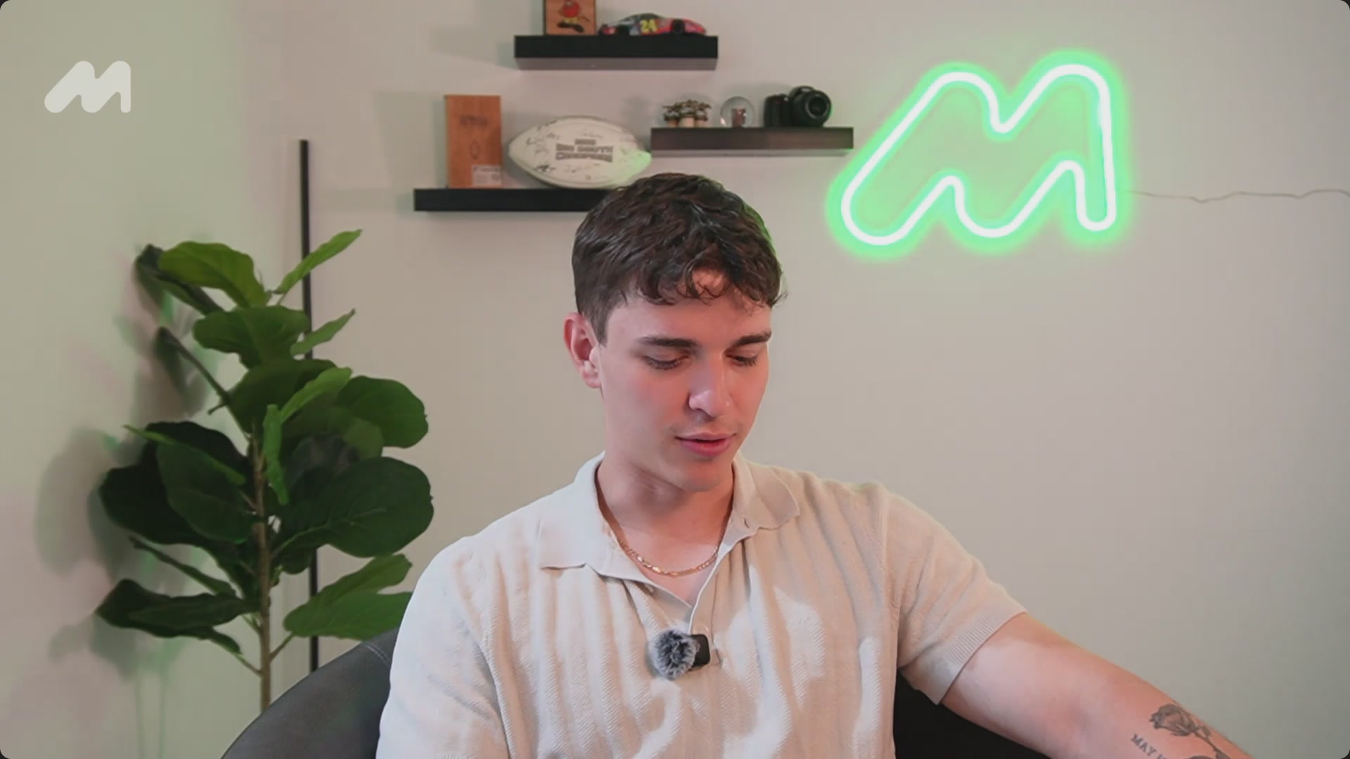 The image features a young man sitting in an office chair with his arm resting on his knee while looking at something off-camera. The background includes a green neon sign that reads "M" above him, suggesting he might be participating in a live stream or video call related to the M logo. There is also a plant and a clock visible in the room, adding some natural elements to the scene.