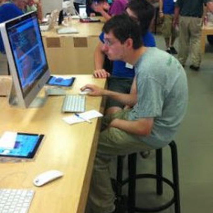 A young man wearing glasses and a tie-dye shirt sits on a stool at an Apple Store, intently using an iMac while a salesperson in a blue shirt leans in to assist him—capturing the moment of a casual shopper buying tech gear after getting swept up in meme-stock hype.