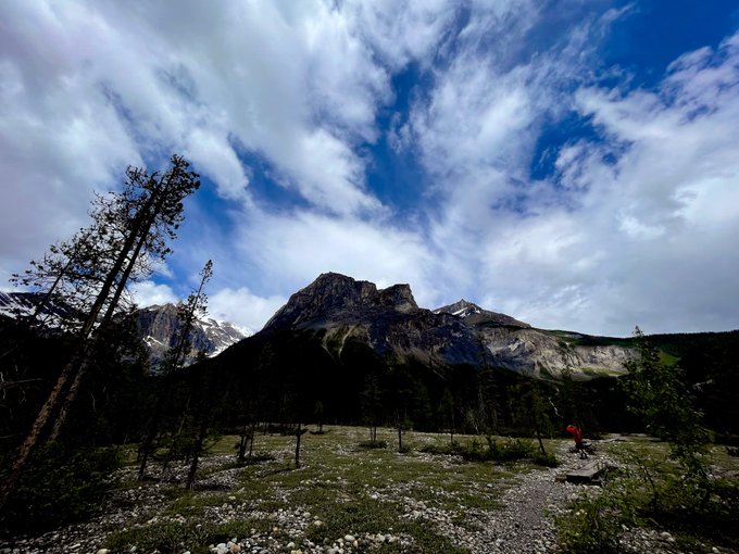 A scenic landscape features a rocky mountain peak rising above a grassy field with scattered trees and a dramatic sky filled with clouds.