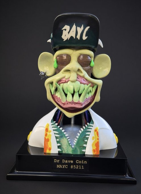 A collectible figurine of a Bored Ape character wearing a 'BAYC' cap and Hawaiian shirt, mounted on a black base with a plaque reading 'Dr Dave Coin MAYC #5211'.
